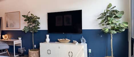 Una sala de estar moderna con un televisor de pantalla plana, un mueble blanco, dos plantas en macetas, un cuadro enmarcado y un sofá gris.
