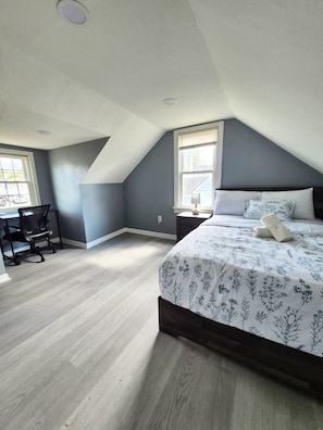 A bedroom with a sloped ceiling, a bed with floral bedding, a desk with a chair, and a nightstand with a lamp.