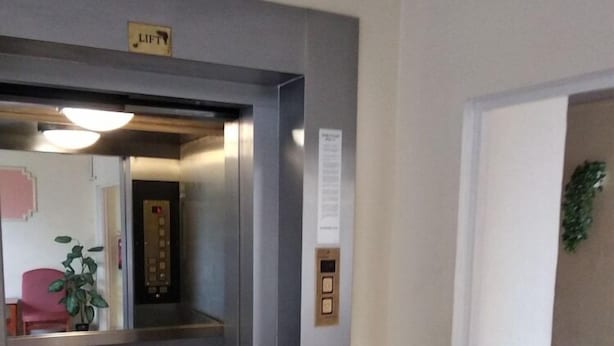 An elevator with a 'LIFT' sign, a wall-mounted information panel, and a potted plant in the background. - Nap (Budapest)