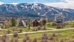 A mountain cabin with a large deck, surrounded by a grassy area and a road.