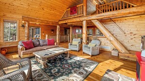 A log cabin interior with a wooden staircase, a sofa set, armchairs, and a patterned rug.