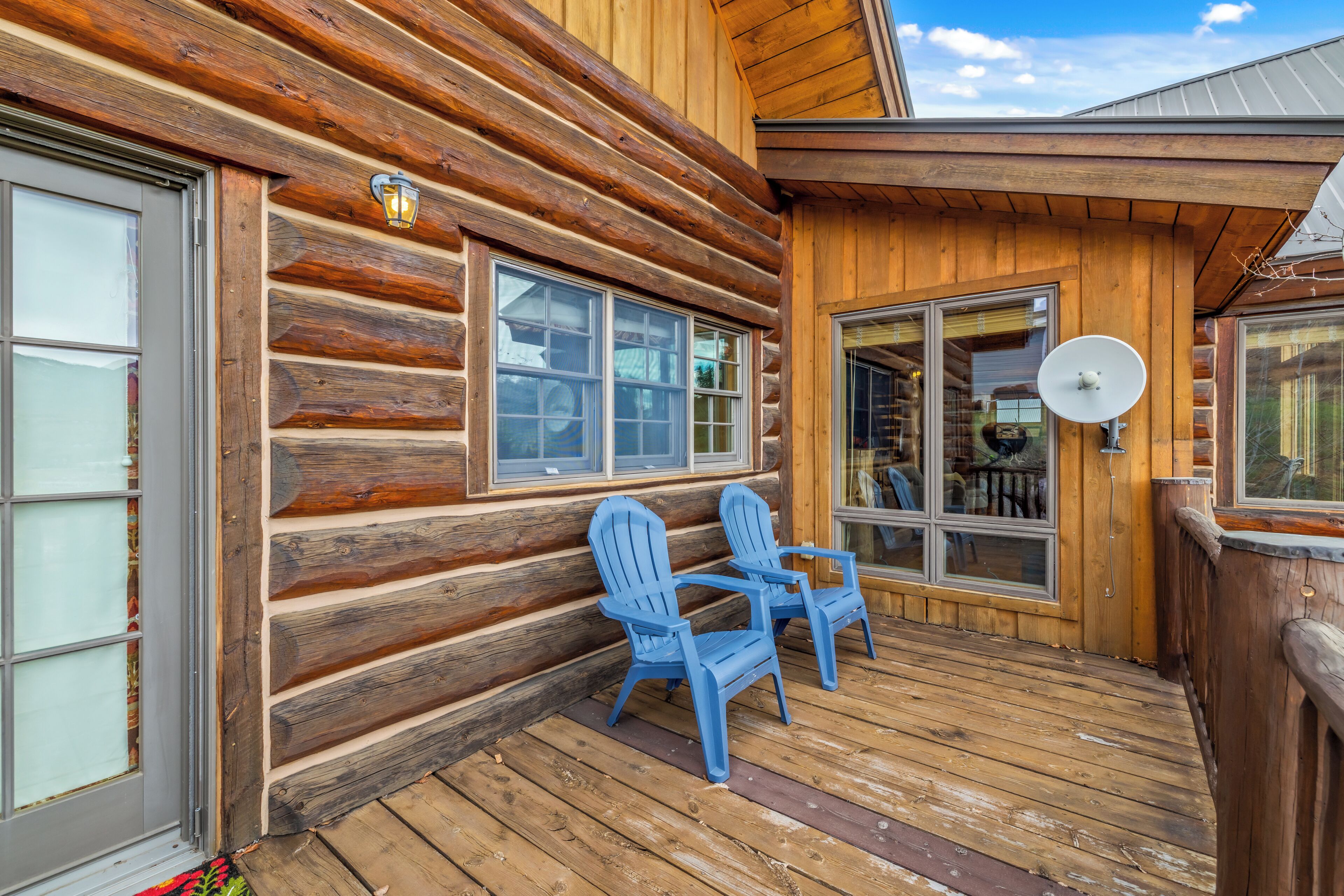 Une cabane en rondins avec une terrasse en bois, deux fauteuils Adirondack bleus et une antenne parabolique.
