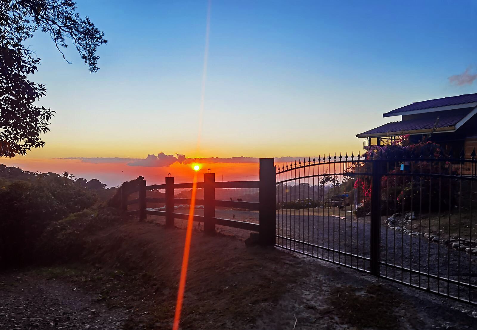 Vista del atardecer desde una zona amurallada con cerca, un edificio con tejado de baldosas y cielo despejado.