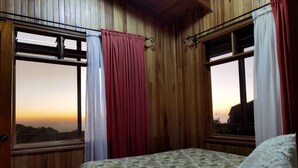A wooden room with a bed, windows with curtains, and a view of the sunset.
