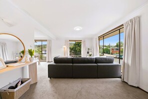 A modern living room with a grey sectional sofa, a round mirror, and a view of greenery outside.