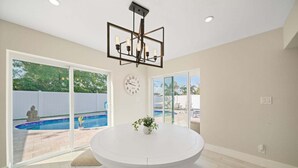 Une pièce avec une table ronde blanche, deux chaises blanches, une horloge au mur et une piscine visible à travers les portes-fenêtres en verre.