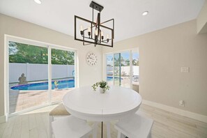 Ein Zimmer mit einem runden weißen Tisch, zwei weißen Stühlen, einer Uhr an der Wand und einem Pool, der durch die Glastüren zu sehen ist.