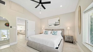 Une chambre avec un grand lit, un ventilateur de plafond, une fenêtre avec des stores et un tableau sur le thème de la plage accroché au mur.