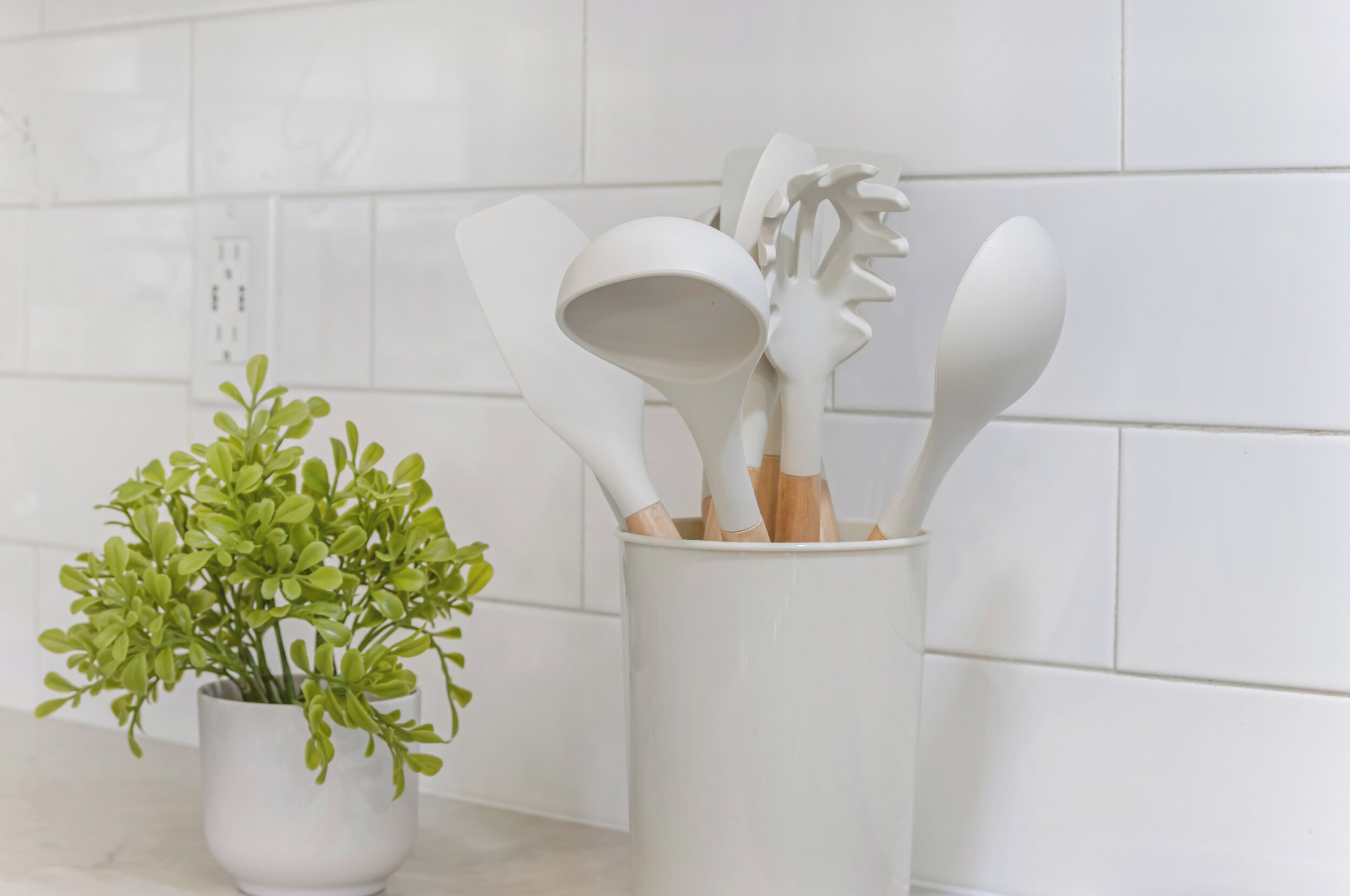 A white ceramic mug with wooden-handled utensils and a green plant on a kitchen counter.