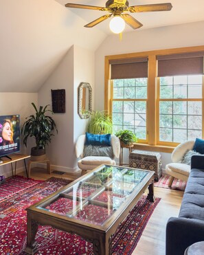 Ein Wohnzimmer mit Deckenventilator, Fernseher, Glastisch, zwei Sofas, einem gemusterten Teppich und einem großen Fenster.