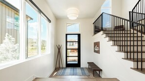 Un couloir doté d’un plancher en bois, d’un banc, d’un tapis, d’une porte donnant sur un beau paysage et d’un escalier avec des rampes noires.