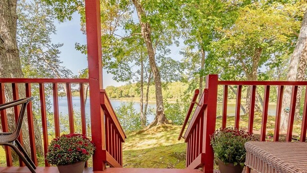 A wooden deck with red railings, a wicker chair, and a table, overlooking a lake with trees in the background.