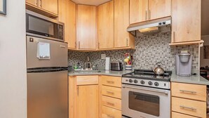 A compact kitchen with wooden cabinets, a stainless steel refrigerator, a microwave, a stove, and a coffee maker.