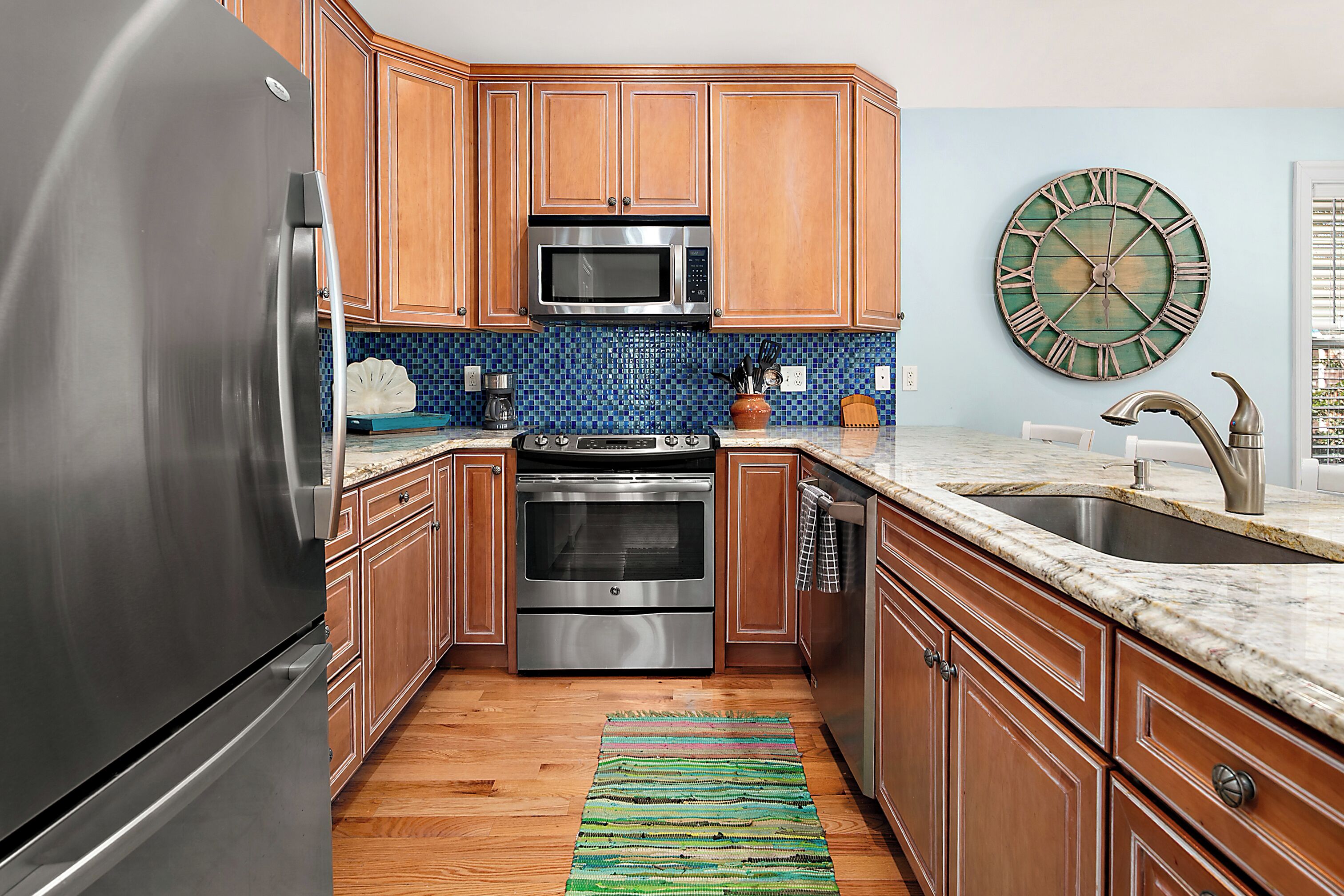 A kitchen with wooden cabinets, stainless steel appliances, a blue tiled backsplash, and a large wall clock.