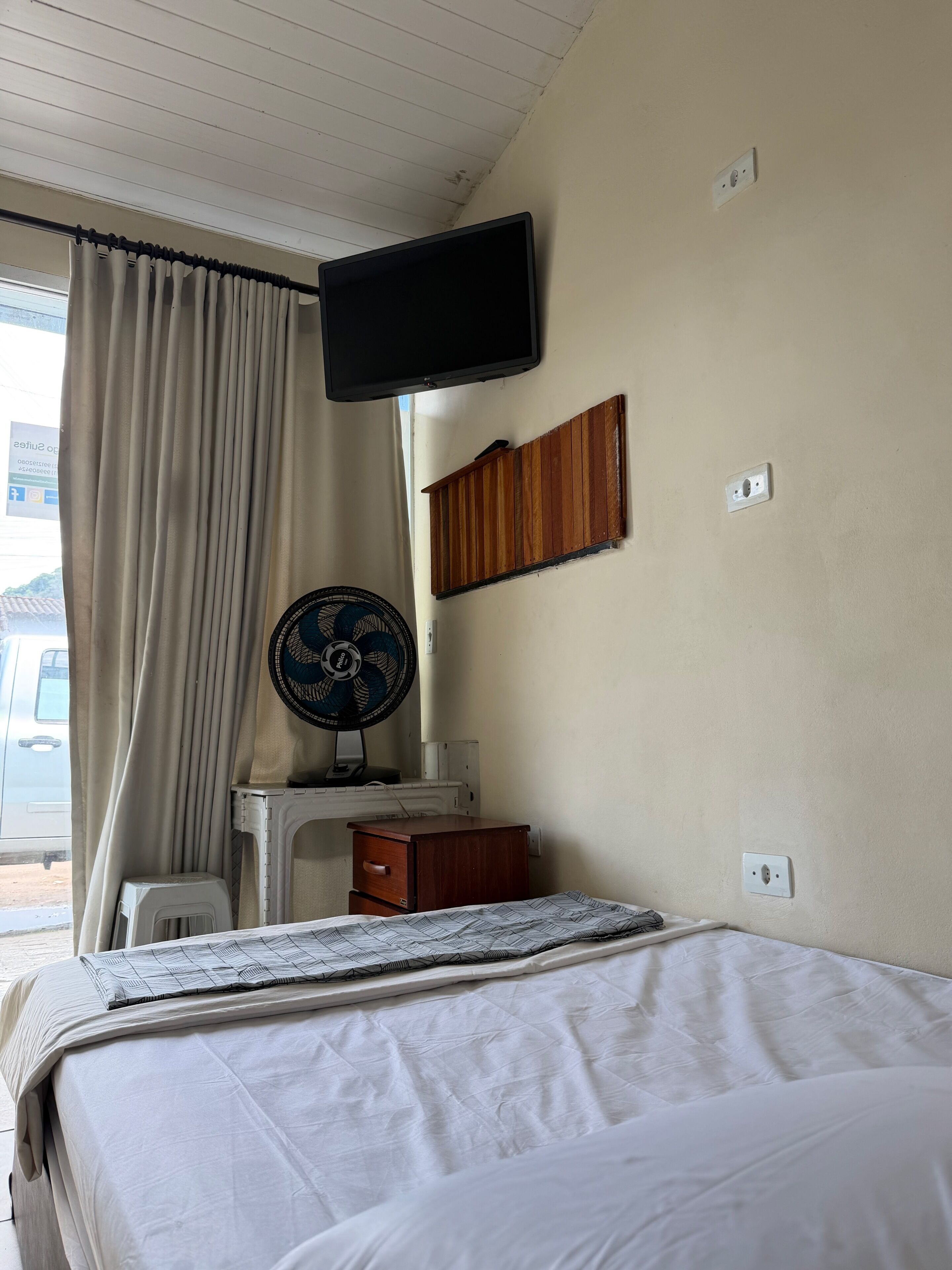 A hotel room with a bed, a TV mounted on the wall, a ceiling fan, a small wooden cabinet, and a window with curtains.