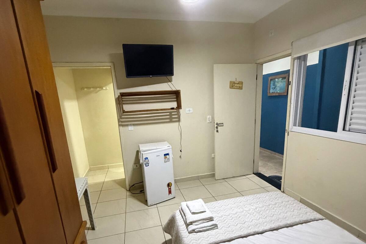 A bedroom with a bed, a television mounted on the wall, a small refrigerator, and a wooden wardrobe.