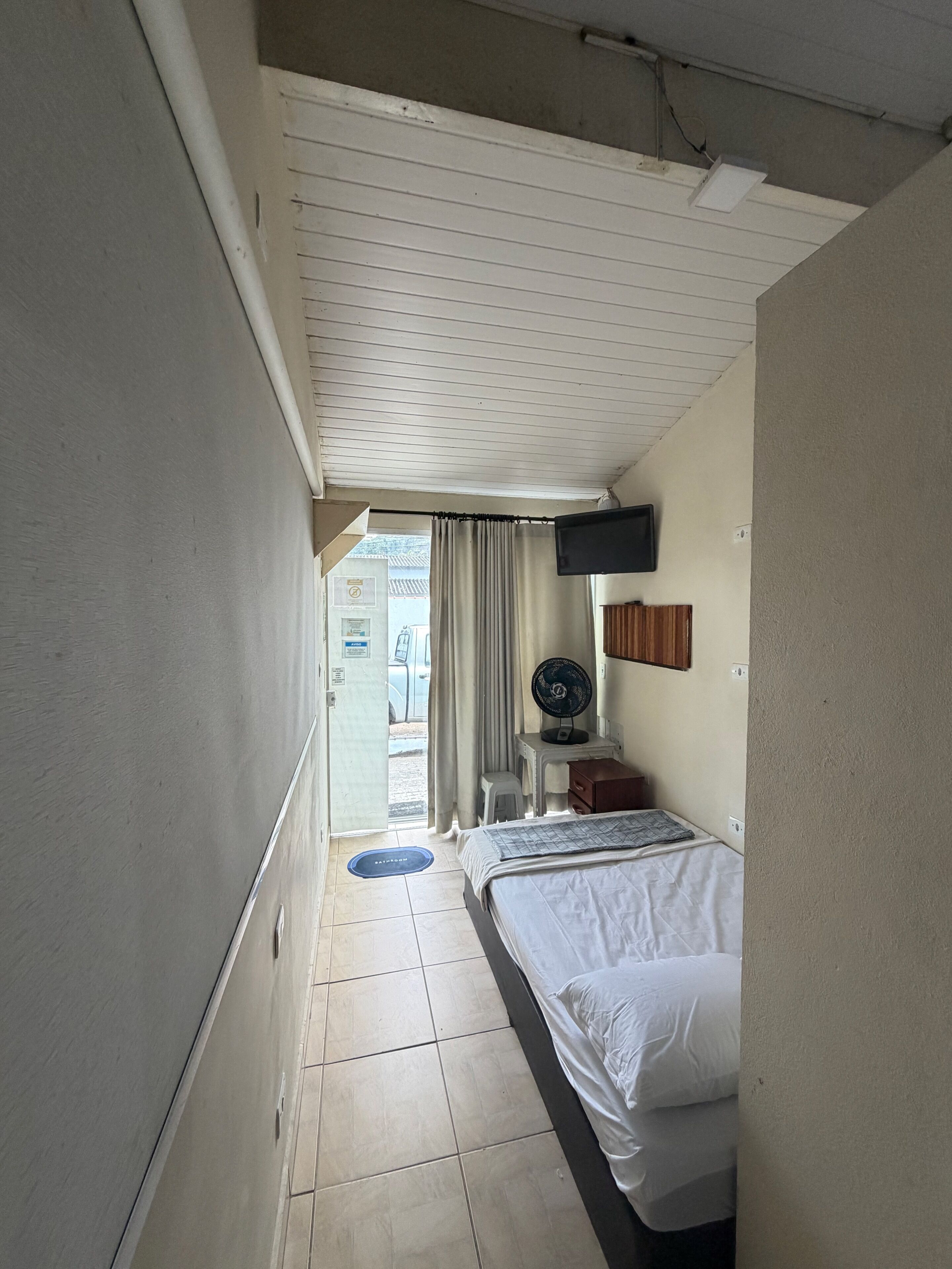 Une chambre d’hôtel étroite, avec un lit, un petit bureau, un téléviseur fixé au mur et une fenêtre avec des rideaux.