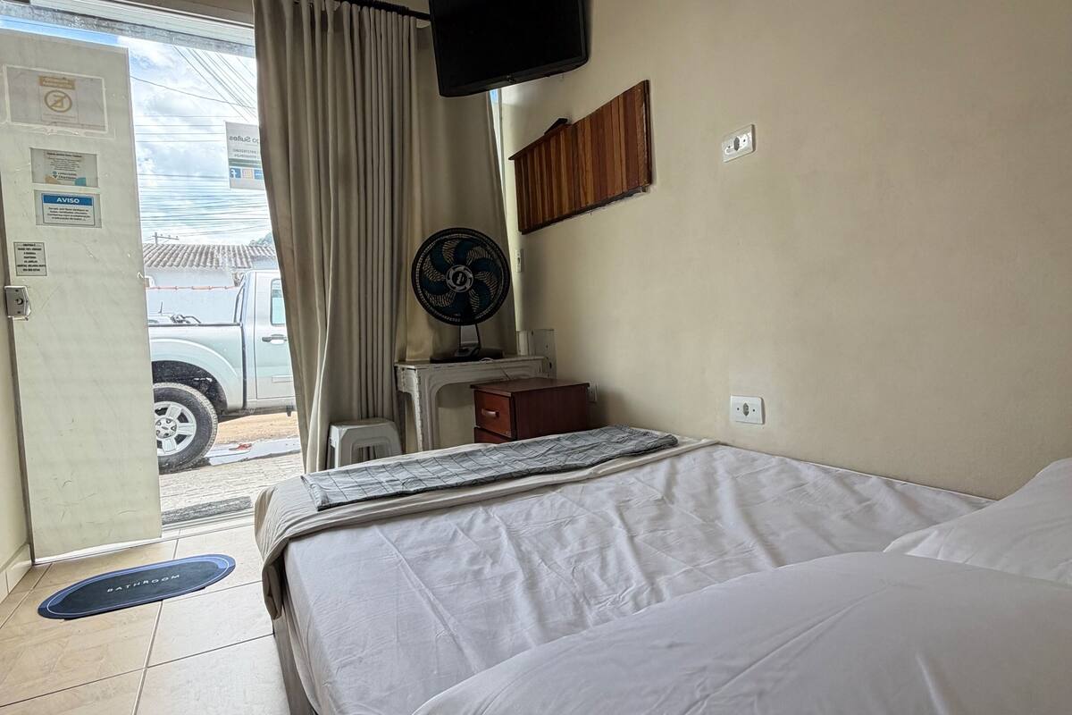 A hotel room with a bed, a television mounted on the wall, a fan, a small wooden cabinet, and a door leading outside.