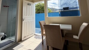 A room with a ceiling fan, a window with a view, a door leading to a shower, and a table with chairs. - Suites Família Otaviano  (Ubatuba)