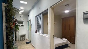 A hallway with a bed, a window with blinds, a small stool, and a plant. - Suites Família Otaviano  (Ubatuba)