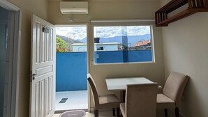A small, well-lit room with a ceiling fan, a table with chairs, a window with a view of a blue wall and mountains, and a door leading outside. - Suites Família Otaviano  (Ubatuba)