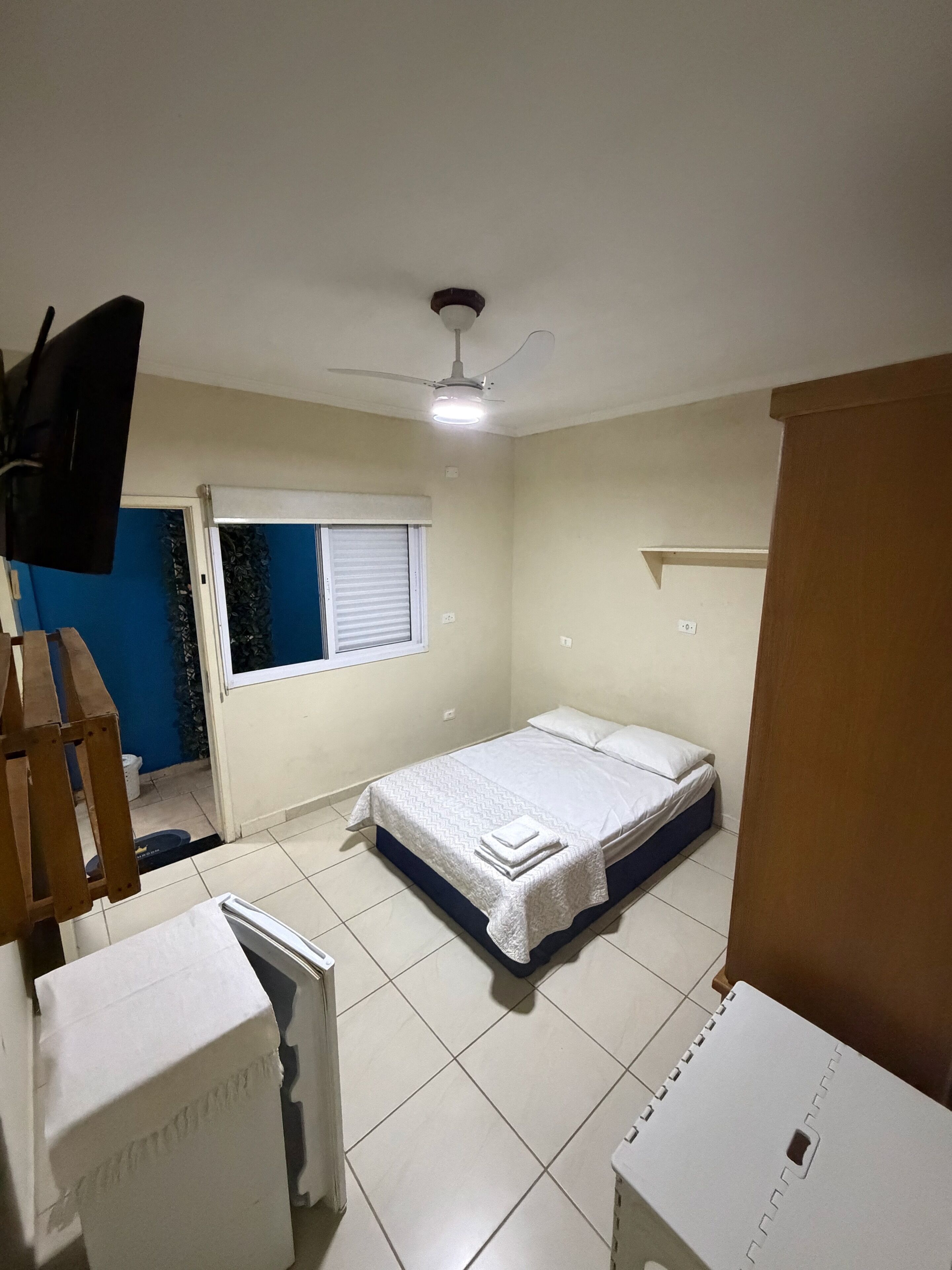 Quarto com cama, ventilador de teto, janela com persianas e televisão.