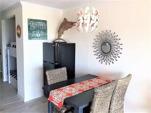 Un coin repas avec une table noire, des coussins à motifs, un chemin de table rouge et blanc, un élément décoratif mural et un réfrigérateur noir.