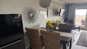 A dining area with wicker chairs, a table with vases of flowers, a wall-mounted TV, and a decorative mirror. - Ocean View Front Row - Cruise Parking - 2 Pools - Hot Tub - GYM -Tennis (Galveston)