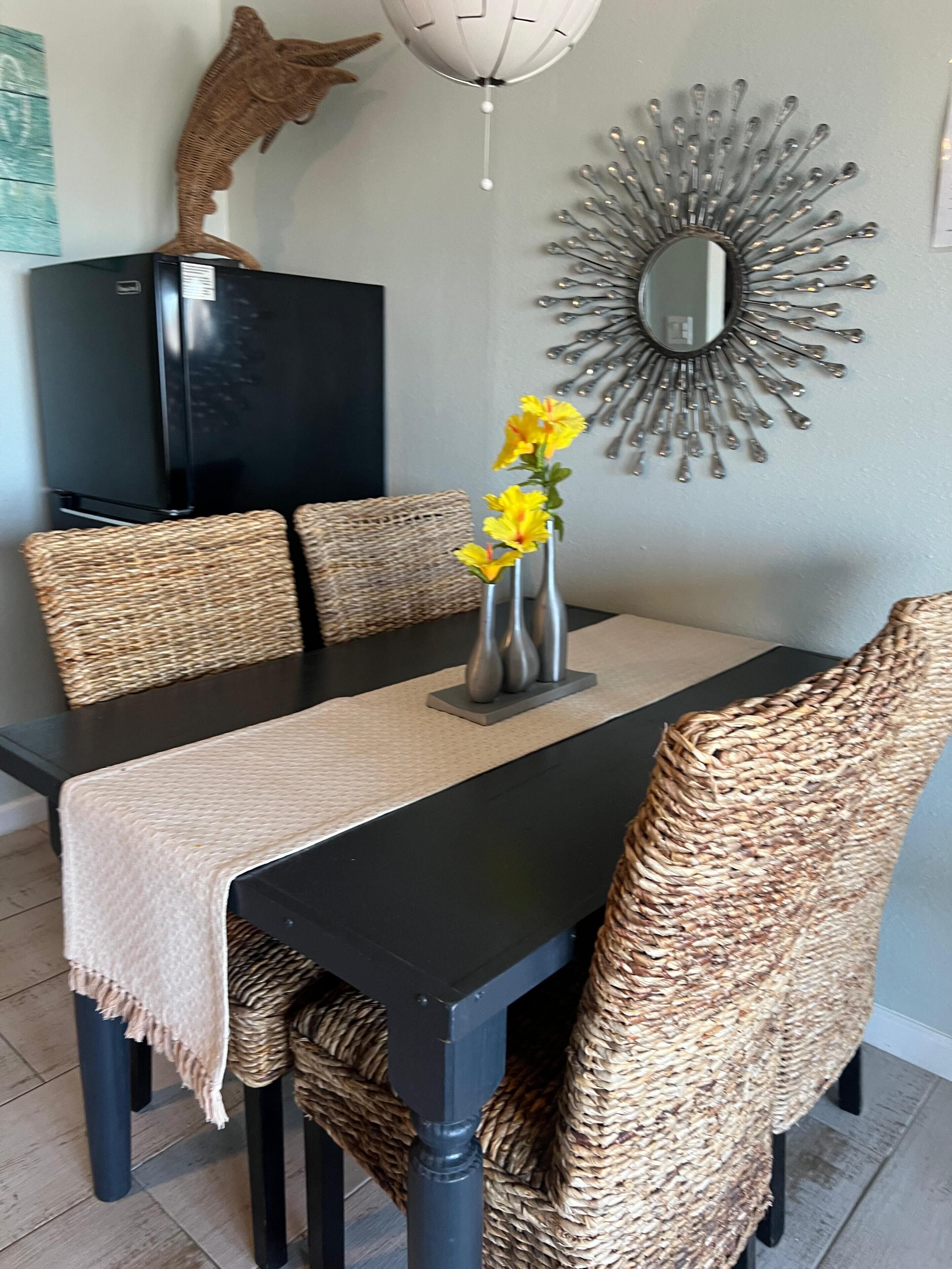 Ein Essbereich mit einem schwarzen Tisch, gewobenen Stühlen, einer Vase mit gelben Blumen, einem dekorativen Spiegel und einer Fischskulptur an der Wand.
