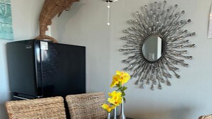 Un comedor con una mesa negra, sillas tejidas, un florero con flores amarillas, un espejo decorativo y una escultura de pez en la pared.