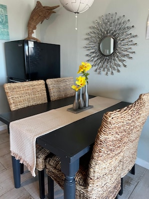 Un comedor con una mesa negra, sillas tejidas, un florero con flores amarillas, un espejo decorativo y una escultura de pez en la pared.