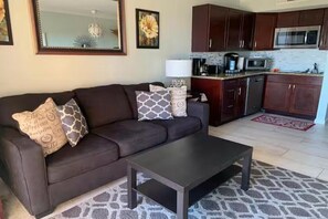 Un salon avec un canapé marron foncé, des coussins à motifs, une table basse, un coin cuisine avec des meubles en bois, un four à micro-ondes et un tapis rouge.