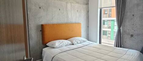 A modern hotel room with a large bed, concrete walls, and a view of the city.