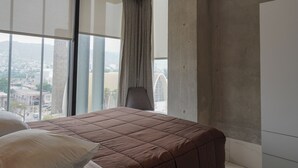 Un dormitorio con un ventanal grande, una cama con colcha marrón, una silla y vista a un paisaje urbano.