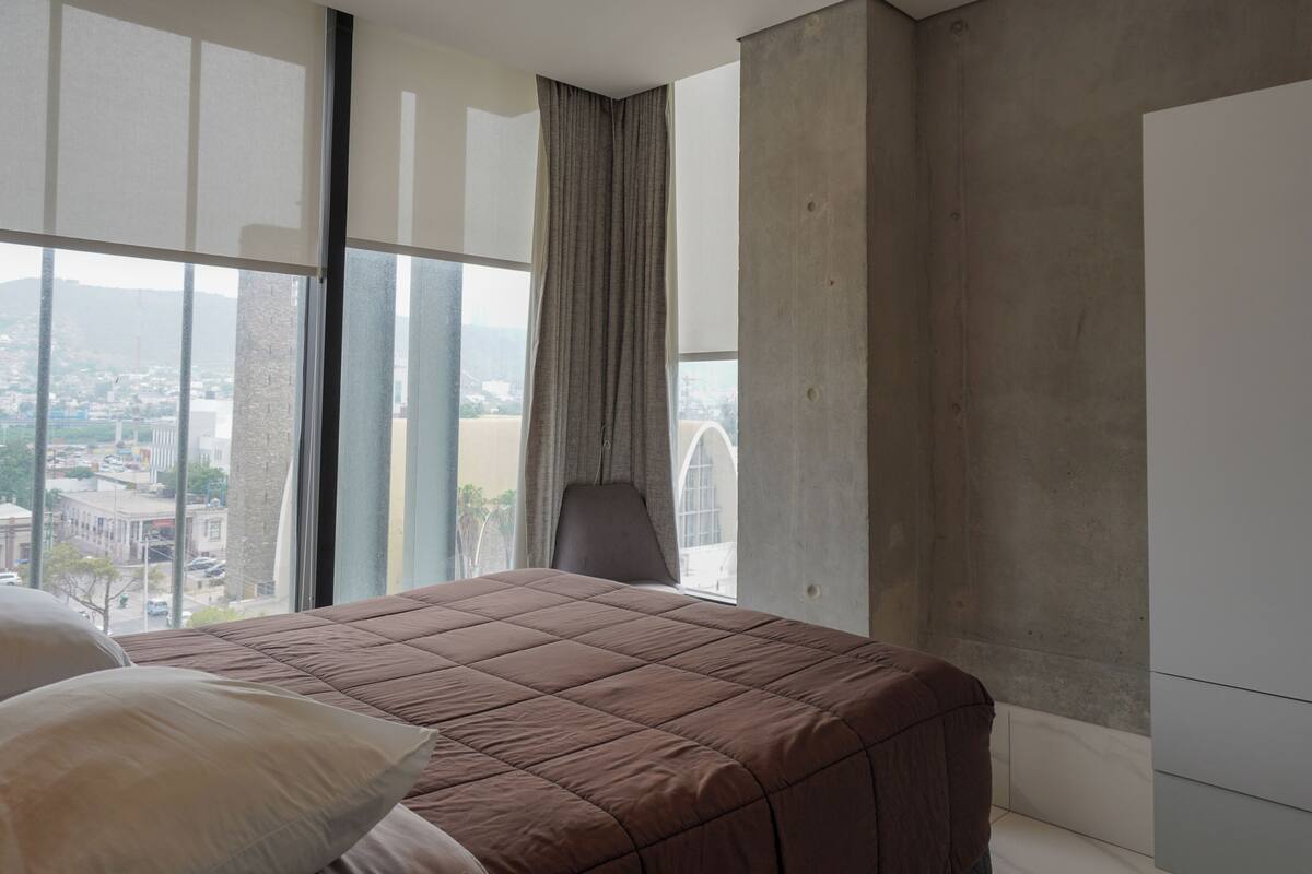 Quarto com uma janela grande, cama com colcha marrom, uma cadeira e vista para a cidade.