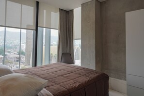 A bedroom with a large window, a bed with a brown quilt, a chair, and a view of a cityscape. - Purísima 5.0 (Monterrey)