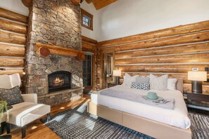 A bedroom with a stone fireplace, wooden beams, a bed with pillows, and a nightstand with a lamp.