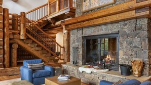 Interior de una cabaña de troncos con chimenea de piedra, asientos tapizados en azul, una escalera de madera y decoración rústica.