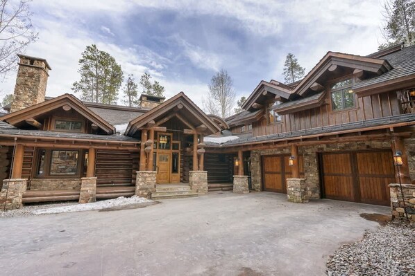 A log cabin with a stone chimney, wooden garage doors, and a gravel driveway.
