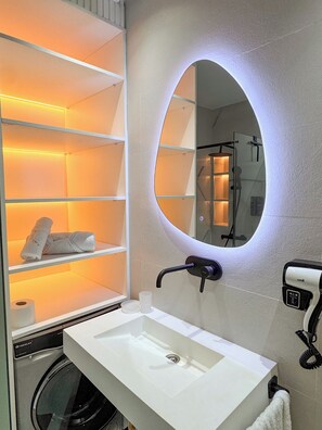 Une salle de bain avec un lavabo blanc, un miroir, des étagères avec des serviettes, un porte-rouleau de papier toilette et un lave-linge.