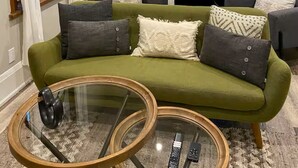 Ein Wohnzimmer mit einem grünen Sofa, einem Glastisch und einem großen Fenster mit Vorhängen.