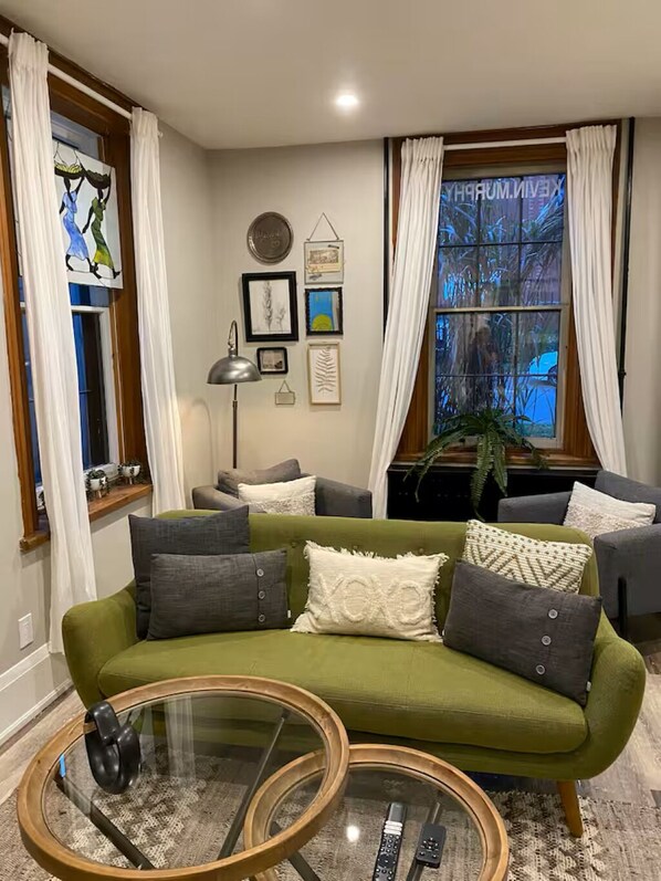 A living room with a green sofa, a glass coffee table, and a large window with curtains.