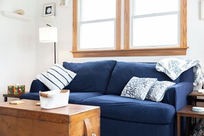 A cozy living room with a blue sofa, a wooden coffee table, and a window with wooden frames.