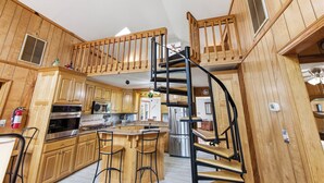 A kitchen with wooden cabinets, a central island with stools, a spiral staircase, and a view of the outside. - Thistle: Semi-Oceanfront home in Duck, Elevator, Sanderling Community Amenities (Duck)