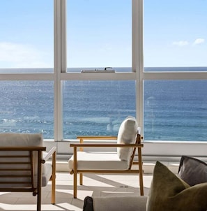Ein Zimmer mit großem Fenster und Meerblick, ein Holzstuhl und eine Couch mit Kissen.