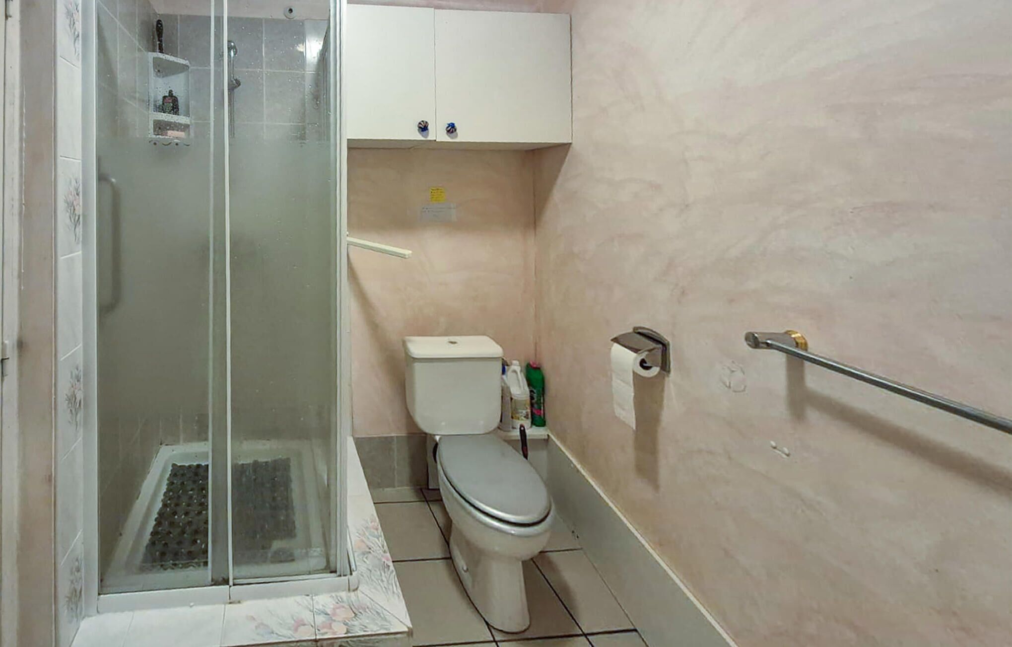 A bathroom with a glass shower enclosure, a toilet, a handrail, and a cabinet above the toilet.