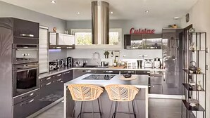 Una cocina moderna con una isla central, dos taburetes de bar, hornos empotrados y una campana extractora de acero inoxidable.