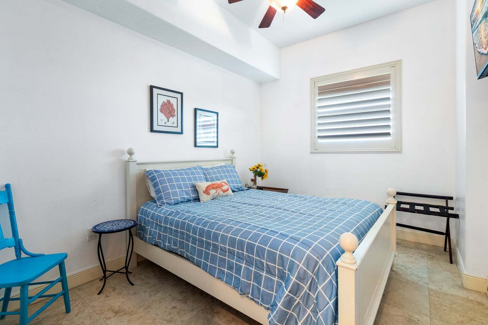 A bedroom with a bed, a chair, a small table, a window with blinds, and two framed pictures on the wall.
