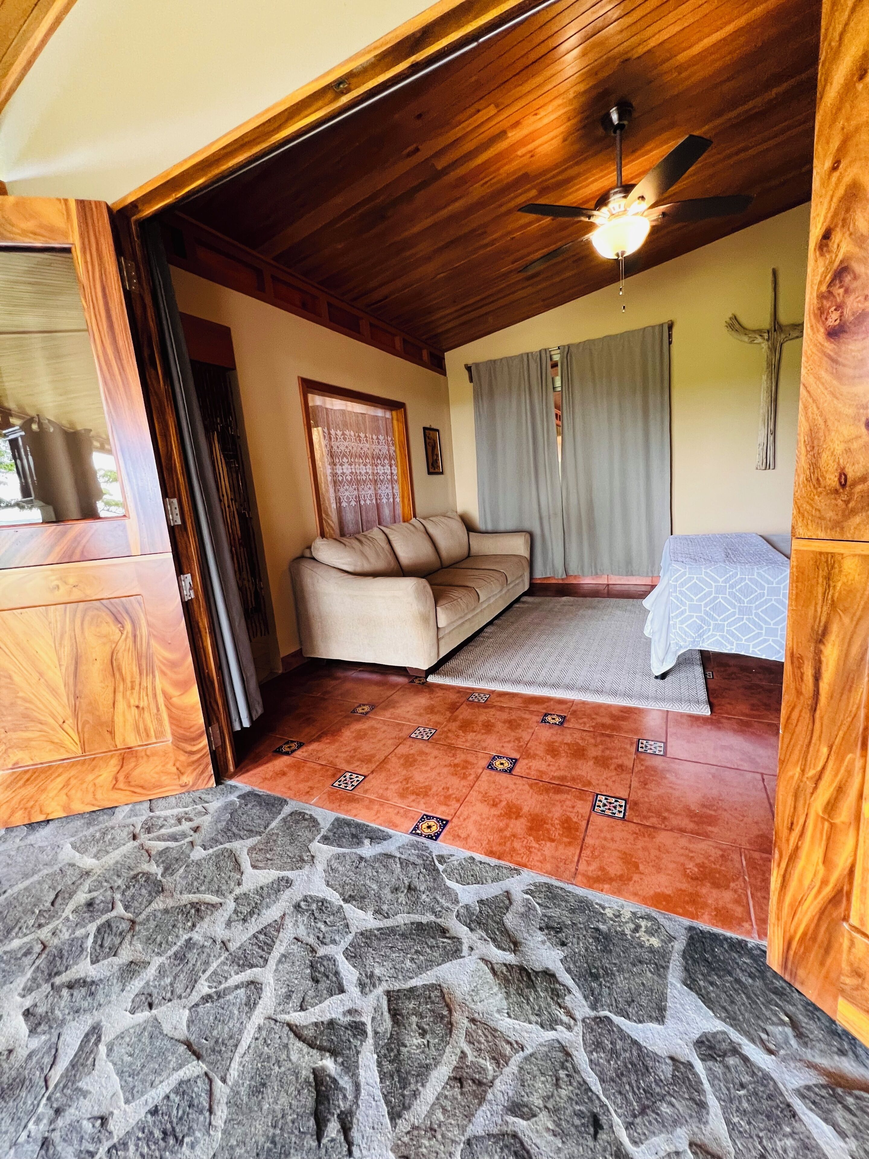 Quarto com soalho de pedra, teto de madeira, ventoinha no teto, sofá, cama e janela com cortinas.