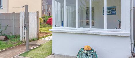 A house with a white exterior, a window with white frames, a small garden with a wooden fence, and a concrete pathway.